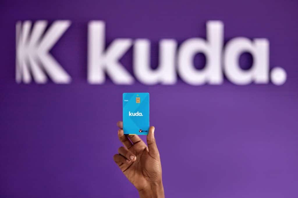 Black hand holding a Kuda card raised high. The background shows Kuda logo and Kuda with a full stop  