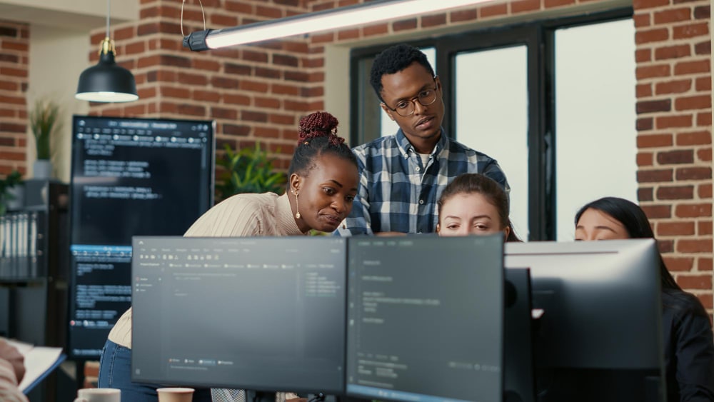 developers brainstorming ideas looking code computer screens asking feedback from senior developer while intern joins discussion junior programmers collaborating group project