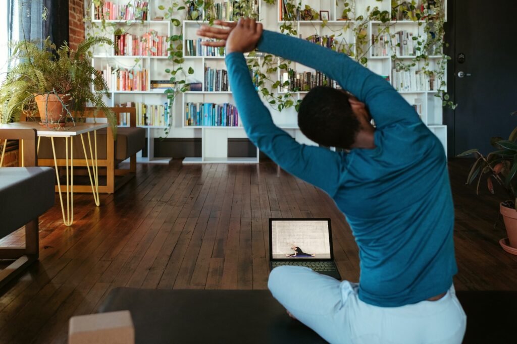 Man sitting on a yoga mat at home stretching with a laptop in front of him