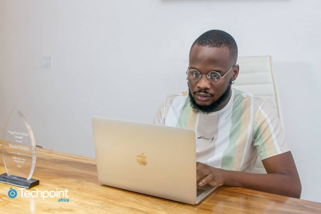 Daniel Orubo. Responsable du contenu et de la stratégie de contenu, Piggyvest assis derrière un bureau et travaillant sur son Mac. Sur le bureau se trouve une plaque de récompense du personnel de l'année PiggyTech Global Limited, 2022.