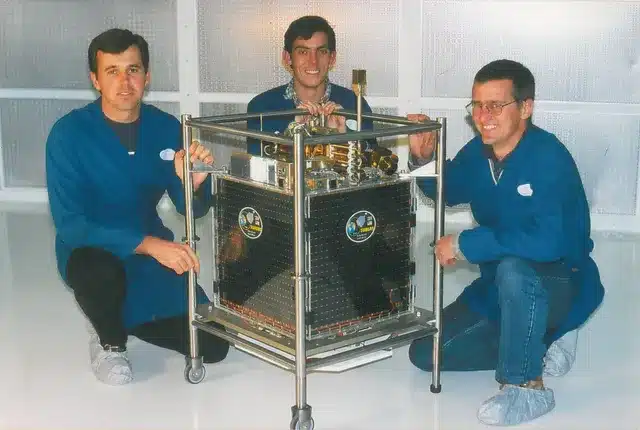 A group of three men kneeling next to what seems to be a satellite device